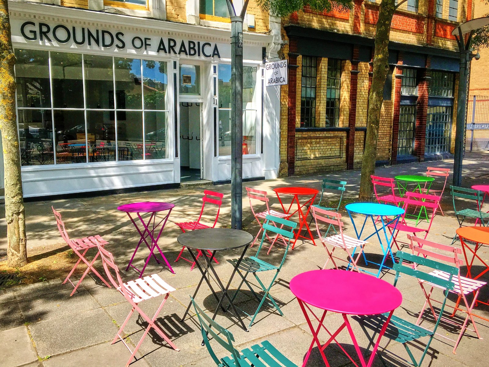 The Grounds of Arabica shopfront on Bramley Road, with brightly coloured tables and chairs outside in the sun